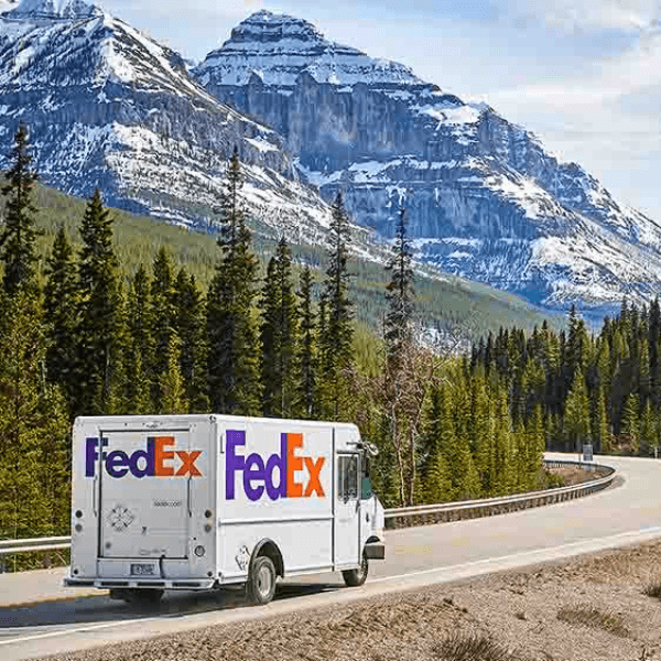 A FedEx delivery truck drives on a winding mountain road lined with evergreen trees, with snow-capped peaks in the background.