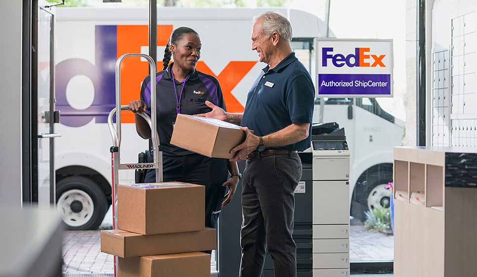A FedEx employee hands a package to a customer inside a FedEx Authorized ShipCenter, with boxes and a delivery truck visible in the background.